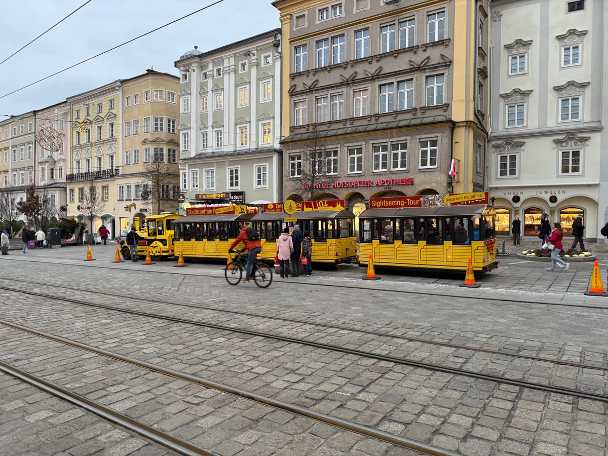 A yellow sightseeing train passes through an unknown historic plaza, filled with festive lights, but no Christmas market decorations.