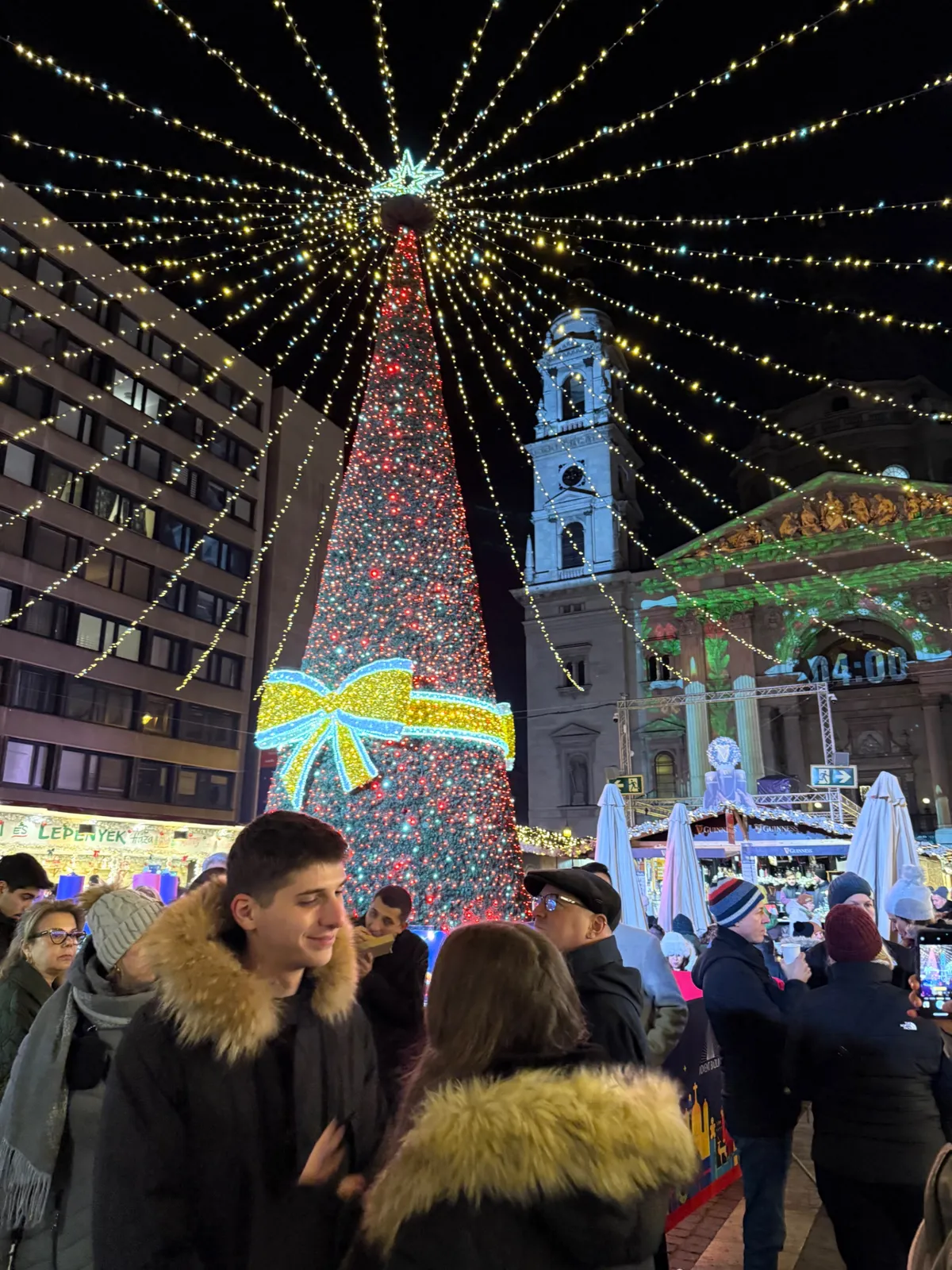 Festive decorations and lights illuminate the Unknown Christmas market with a red tree and joyful crowds.